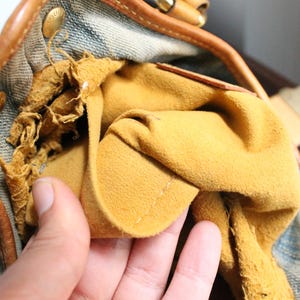 May include: A handbag interior featuring a mustard yellow suede lining and a blue denim exterior. The leather trim is tan with gold-tone metal hardware. The suede lining shows signs of wear and fraying along the upper edge near the leather piping.