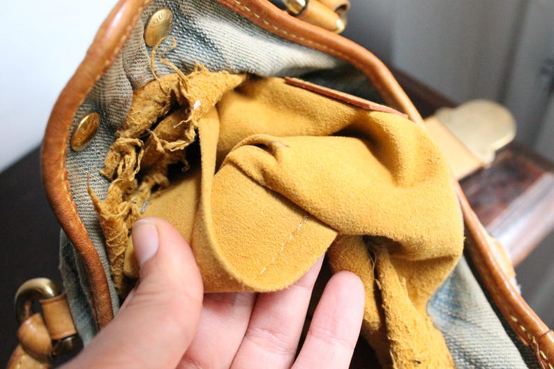 May include: A handbag interior featuring a mustard yellow suede lining and a blue denim exterior. The leather trim is tan with gold-tone metal hardware. The suede lining shows signs of wear and fraying along the upper edge near the leather piping.
