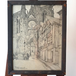 May include: A framed black and white sketch of a European street scene, featuring a large cathedral and buildings with detailed architectural elements. The artwork is displayed on a wooden easel, showcasing the artist's rendering of the city.
