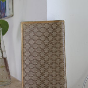 K&ouml;nnte beinhalten: Eine braune und beigefarbene Brieftasche mit einem sich wiederholenden Muster des Buchstabens "C" und einem kleineren Muster eines Quadrats. Die Brieftasche hat einen braunen Lederbesatz.