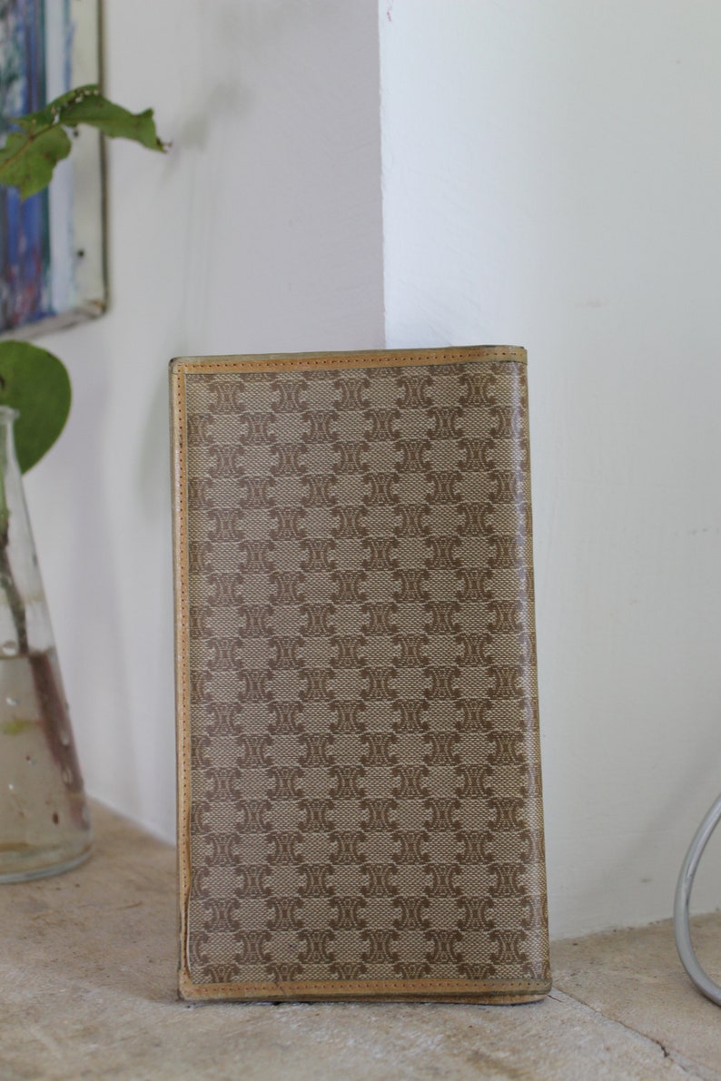 K&ouml;nnte beinhalten: Eine braune und beigefarbene Brieftasche mit einem sich wiederholenden Muster des Buchstabens "C" und einem kleineren Muster eines Quadrats. Die Brieftasche hat einen braunen Lederbesatz.