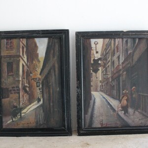 Peut inclure: Deux peintures encadrées représentant des scènes de rue dans une ville européenne. Les deux peintures présentent des rues étroites et pavées bordées de bâtiments. La peinture de gauche montre une rue avec une charrette et une personne qui marche. La peinture de droite montre une rue avec une personne qui marche et une enseigne suspendue au-dessus de la rue.