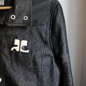 Puede incluir: Una chaqueta de cuero negro con un acabado texturizado y desgastado, que presenta un logotipo blanco en el pecho, un cuello puntiagudo con dos ojales met&aacute;licos plateados y un cierre frontal de botones. La chaqueta cuelga de una percha de madera.