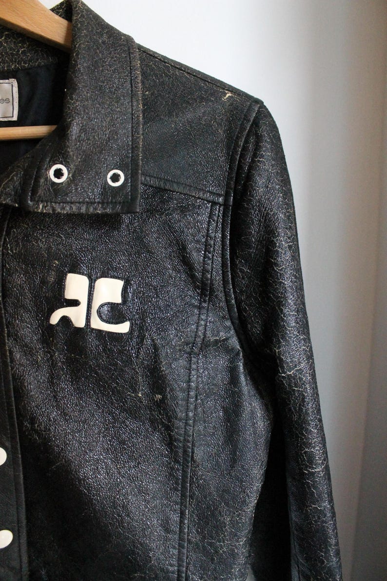 Puede incluir: Una chaqueta de cuero negro con un acabado texturizado y desgastado, que presenta un logotipo blanco en el pecho, un cuello puntiagudo con dos ojales met&aacute;licos plateados y un cierre frontal de botones. La chaqueta cuelga de una percha de madera.