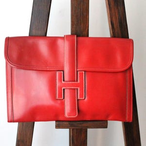 May include: A red leather clutch bag with a large, white, stitched H-shaped closure.