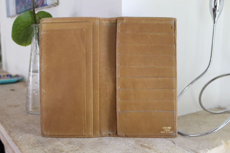 K&ouml;nnte beinhalten: Eine braune Ledergeldb&ouml;rse mit mehreren Kartenf&auml;chern und einem Geldscheinfach. Die Geldb&ouml;rse ist ge&ouml;ffnet und das Innere ist sichtbar. Der Markenname "CELINE PARIS" ist in Gold auf der Innenseite der Geldb&ouml;rse aufgedruckt.