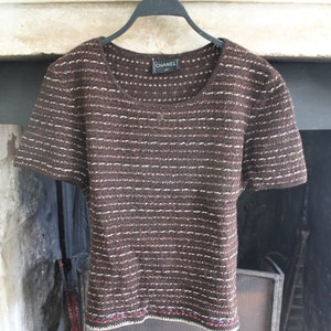 May include: A brown and white striped short-sleeved top with a black label that says "CHANEL" on the front. The top has a colourful embroidered trim around the bottom.