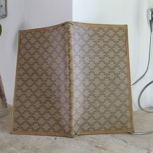 K&ouml;nnte beinhalten: Eine braune Ledergeldb&ouml;rse mit einem sich wiederholenden Muster des Wortes "CELINE" in einer kleineren Schriftart. Die Geldb&ouml;rse ist geschlossen und liegt auf einer hellbraunen Oberfl&auml;che.