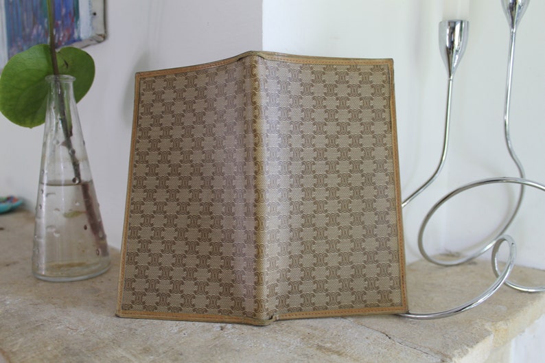 K&ouml;nnte beinhalten: Eine braune Ledergeldb&ouml;rse mit einem sich wiederholenden Muster des Wortes "CELINE" in einer kleineren Schriftart. Die Geldb&ouml;rse ist geschlossen und liegt auf einer hellbraunen Oberfl&auml;che.