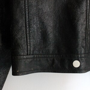 Puede incluir: Una chaqueta de cuero negro con una superficie texturizada y una tapeta frontal con botones de presi&oacute;n circulares blancos. La prenda incluye detalles de costuras verticales en el panel frontal y un dobladillo recto con costuras visibles.