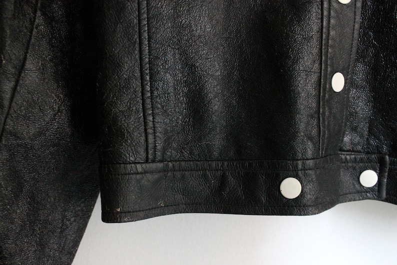Puede incluir: Una chaqueta de cuero negro con una superficie texturizada y una tapeta frontal con botones de presi&oacute;n circulares blancos. La prenda incluye detalles de costuras verticales en el panel frontal y un dobladillo recto con costuras visibles.
