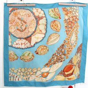 May include: A square silk scarf with a light blue background featuring a large circular arrangement of various seashells and a central anchor motif, surrounded by scattered individual shells and a cascading pattern of brown and white seashells.