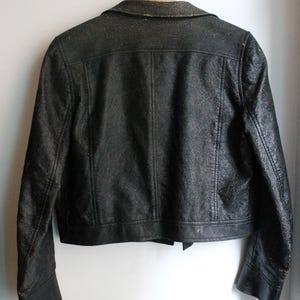 Puede incluir: Una chaqueta negra con un acabado texturizado y desgastado, con cuello, colgada de una percha de madera. La chaqueta presenta mangas largas con pu&ntilde;os, un dobladillo recto y detalles de costuras verticales en la espalda y los hombros.
