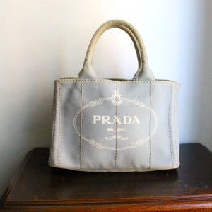 Peut inclure: Un sac fourre-tout en toile bleu clair doté de deux poignées supérieures et de détails surpiqués verticaux. L'avant présente un logo ovale brodé en blanc avec le texte PRADA MILANO centré, entouré d'une bordure en forme de corde.