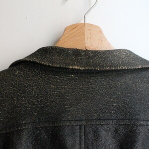 Puede incluir: Una chaqueta de cuero negro con un acabado texturizado y desgastado, con signos visibles de uso a lo largo del cuello y las costuras de los hombros. La chaqueta presenta un dise&ntilde;o de cuello cl&aacute;sico y costuras visibles en el panel trasero.