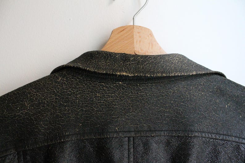 Puede incluir: Una chaqueta de cuero negro con un acabado texturizado y desgastado, con signos visibles de uso a lo largo del cuello y las costuras de los hombros. La chaqueta presenta un dise&ntilde;o de cuello cl&aacute;sico y costuras visibles en el panel trasero.