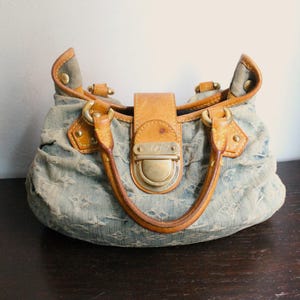 May include: A blue denim handbag featuring a monogram pattern, tan leather trim, and gold-tone hardware. The bag has two rounded leather handles, a central leather strap with a metallic push-lock closure, and gathered fabric detailing.