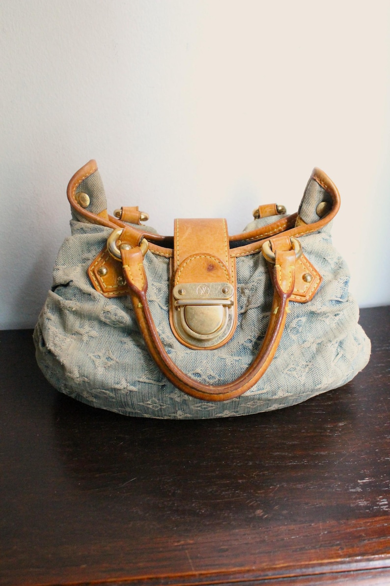 May include: A blue denim handbag featuring a monogram pattern, tan leather trim, and gold-tone hardware. The bag has two rounded leather handles, a central leather strap with a metallic push-lock closure, and gathered fabric detailing.