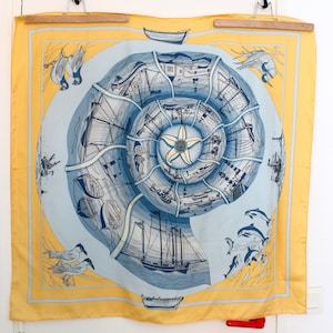 May include: A square silk scarf featuring a yellow border and a central blue and white spiral shell design. The shell contains illustrations of various sailing ships, lighthouses, and nautical scenes, with seagulls and dolphins in the corners.