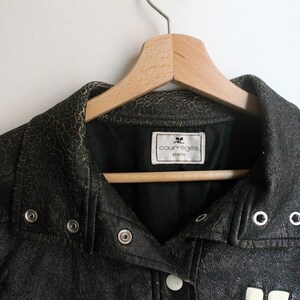 Puede incluir: Una chaqueta de cuero negro con una textura agrietada y desgastada, que presenta un cuello puntiagudo, botones de presi&oacute;n de metal plateado y una etiqueta interior blanca que dice Courr&egrave;ges Paris. La chaqueta est&aacute; en una percha de madera.
