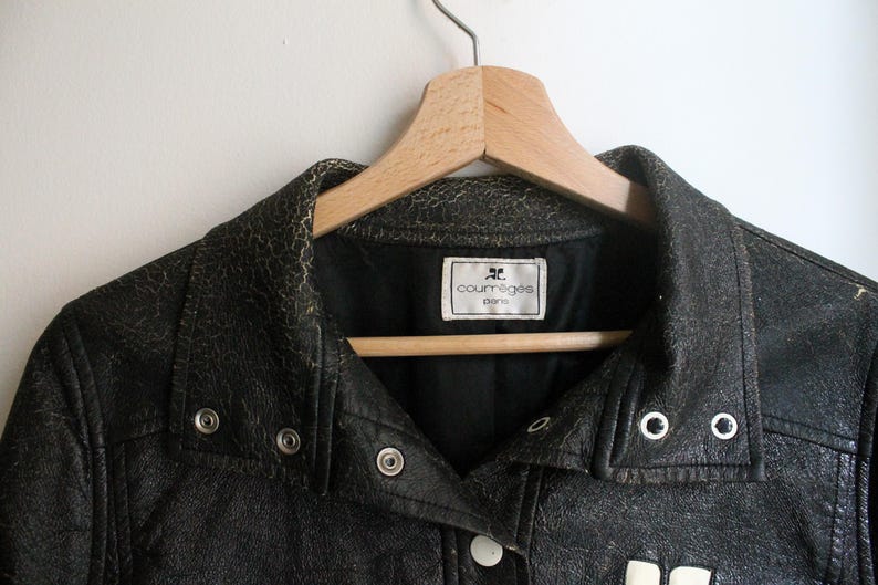 Puede incluir: Una chaqueta de cuero negro con una textura agrietada y desgastada, que presenta un cuello puntiagudo, botones de presi&oacute;n de metal plateado y una etiqueta interior blanca que dice Courr&egrave;ges Paris. La chaqueta est&aacute; en una percha de madera.