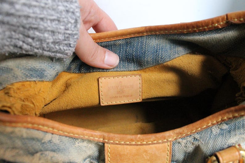 May include: A blue denim handbag with tan leather trim and a mustard yellow interior lining. A rectangular tan leather patch is stitched inside, embossed with the text LOUIS VUITTON PARIS MADE IN FRANCE. The bag features a monogram pattern.