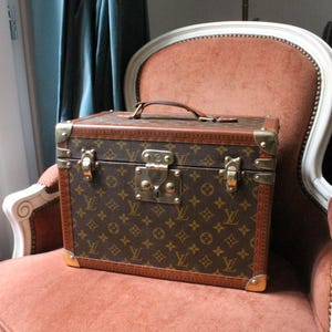 Puede incluir: Baúl de viaje vintage marrón y beige con asa, herrajes de latón y un patrón de logotipo repetido. El baúl rectangular está sobre una silla tapizada rosa. El baúl tiene un asa de cuero y protectores de esquina de latón.
