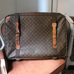 Könnte beinhalten: Braune und beige gemusterte Reisetasche mit Lederriemen und Schnallen. Der rechteckige Koffer steht auf einem pfirsichfarbenen Stuhl. Der Koffer hat einen Griff oben und ist ein klassisches Reiseaccessoire.