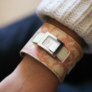 May include: A silver watch with a white face and a pink and white patterned leather strap. The watch face has the text "Christian Dior" printed on it.
