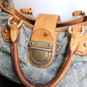 May include: A light blue denim handbag featuring a repeating monogram pattern, tan leather trim, and gold-tone hardware. The bag includes two leather handles and a central front flap closure with a metallic clasp embossed with a logo.