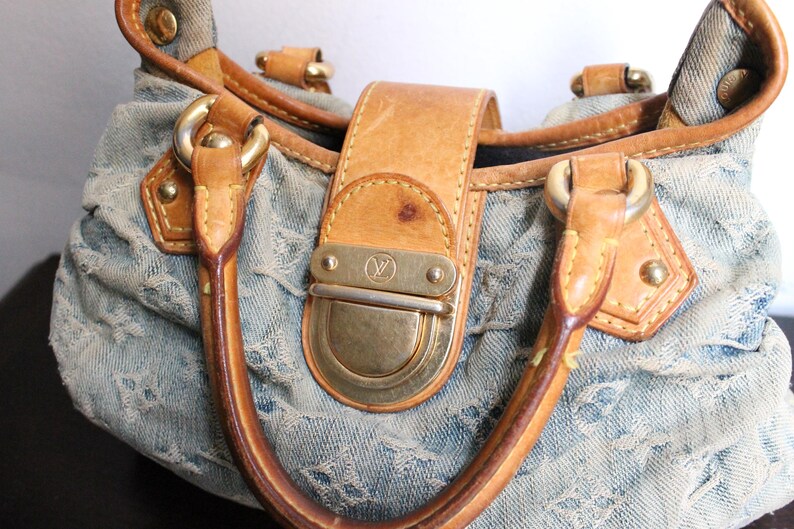 May include: A light blue denim handbag featuring a repeating monogram pattern, tan leather trim, and gold-tone hardware. The bag includes two leather handles and a central front flap closure with a metallic clasp embossed with a logo.