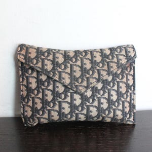 May include: A rectangular envelope-style clutch bag featuring a woven beige and dark gray monogram pattern. The exterior displays a repeating Dior logo print across the fabric surface, with a pointed flap closure and structured edges.