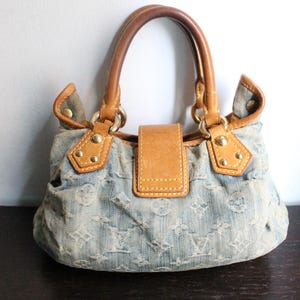 May include: A light blue denim handbag featuring a repeating monogram pattern, tan leather trim, two rounded leather handles, and a central leather flap closure secured with gold-tone metal hardware and rivets on the front panel.