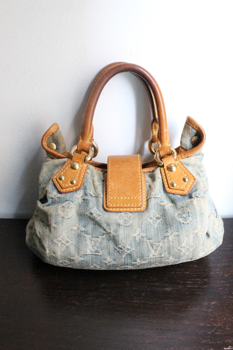 May include: A light blue denim handbag featuring a repeating monogram pattern, tan leather trim, two rounded leather handles, and a central leather flap closure secured with gold-tone metal hardware and rivets on the front panel.
