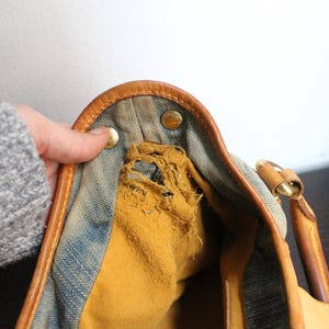 May include: A blue denim handbag featuring tan leather trim and gold-tone metal hardware. The interior lining is a mustard yellow fabric that shows significant fraying and wear along the upper edge near the metal snap closures.