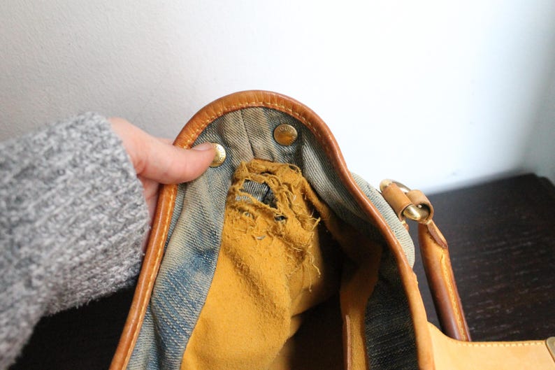 May include: A blue denim handbag featuring tan leather trim and gold-tone metal hardware. The interior lining is a mustard yellow fabric that shows significant fraying and wear along the upper edge near the metal snap closures.