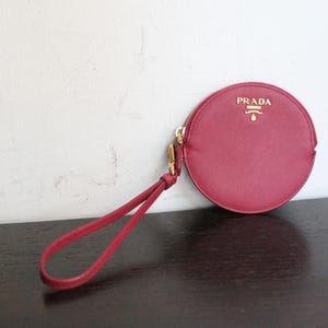Peut inclure: Un porte-monnaie circulaire rose foncé à la finition texturée, doté d'une fermeture éclair dorée et d'une dragonne assortie. L'avant présente le logo et l'emblème Prada dorés. L'accessoire possède une silhouette ronde et compacte.