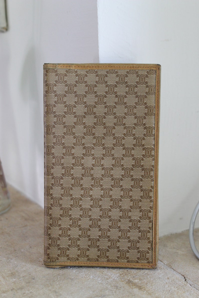 K&ouml;nnte beinhalten: Eine braune Ledergeldb&ouml;rse mit einer beigefarbenen Stoffvorderseite, die ein sich wiederholendes Muster aus den Buchstaben "C" und "E" aufweist.