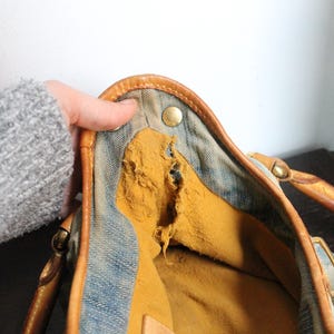 May include: A blue denim handbag featuring tan leather trim and handles with brass-toned metal hardware. The interior is lined with a mustard yellow fabric that shows significant wear, including a large tear along the side seam.