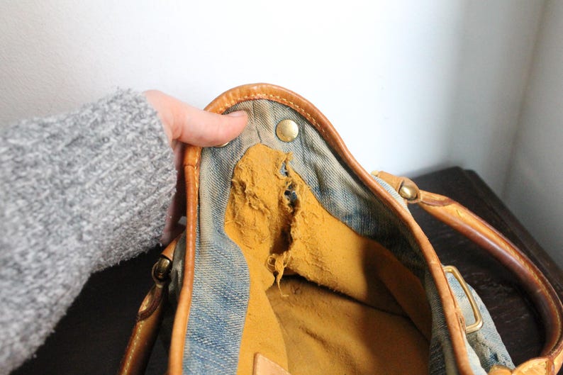 May include: A blue denim handbag featuring tan leather trim and handles with brass-toned metal hardware. The interior is lined with a mustard yellow fabric that shows significant wear, including a large tear along the side seam.