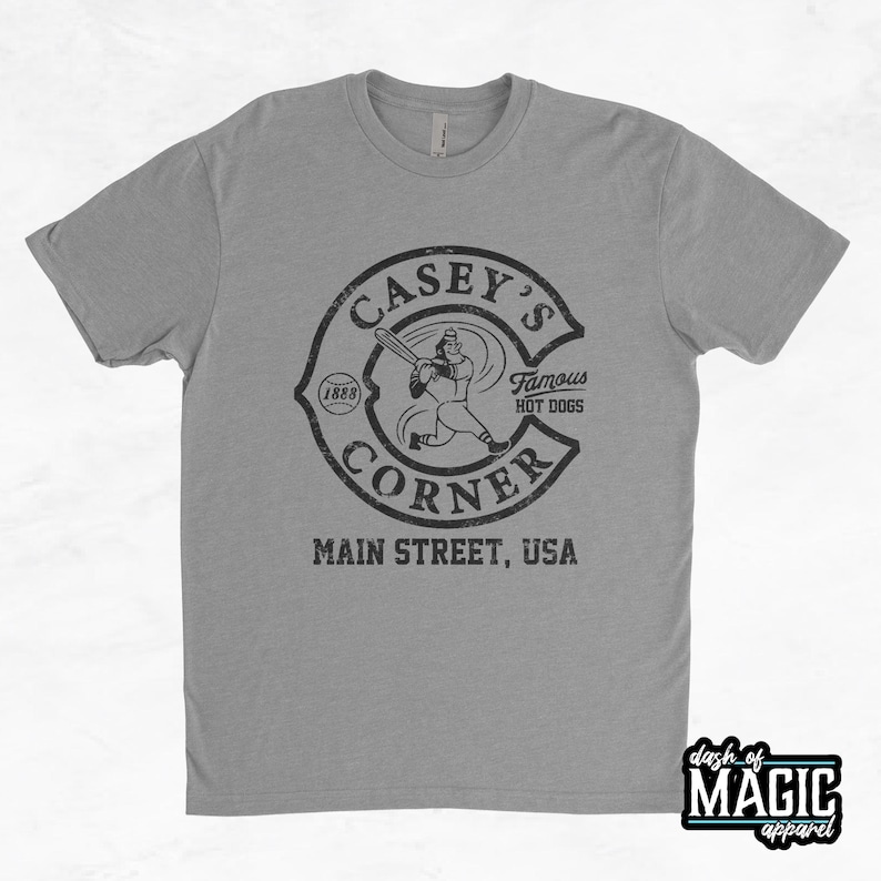 K&ouml;nnte beinhalten: Ein graues T-Shirt mit einem Vintage-Logo f&uuml;r Casey's Corner. Das Logo zeigt einen Baseballspieler in einem Kreis mit dem Text "Casey's Corner" und "Famous Hot Dogs" in schwarzer Schrift. Der Text "Main Street, USA" befindet sich unter dem Logo.