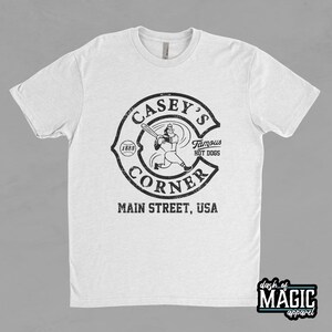 K&ouml;nnte beinhalten: Wei&szlig;es T-Shirt mit einer schwarz-wei&szlig;en Grafik eines Baseballspielers und dem Text "Casey's Corner, Main Street, USA, Famous Hot Dogs, (1888)"