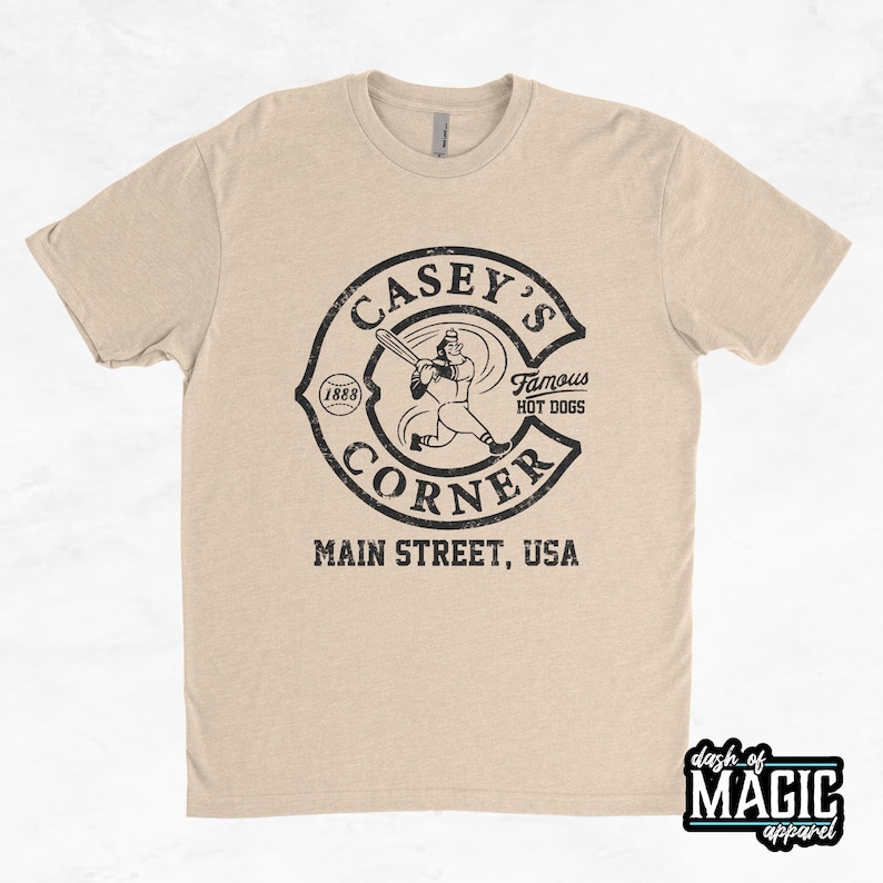 K&ouml;nnte beinhalten: Ein beiges T-Shirt mit einer schwarz-wei&szlig;en Grafik eines Baseballspielers und dem Text "Casey's Corner, Famous Hot Dogs, Main Street, USA".