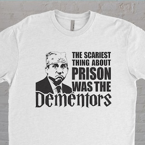 May include: White t-shirt with a black and white graphic of a man wearing a prison hat. The text on the shirt reads "The scariest thing about prison was the Dementors."