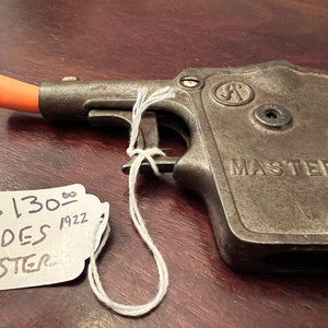 Op de afbeelding: Een vintage metalen speelgoedpistool met een oranje plastic loop. Het pistool is gemarkeerd met "MASTER" en heeft een prijskaartje met de tekst "$130.00 ANDES 1922 MASTER".