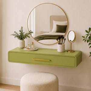 Op de afbeelding: Een aan de muur gemonteerde zwevende kaptafel in een matte olijfgroene afwerking met één lade en een slanke gouden handgreep. Boven de kaptafel hangt een grote ronde spiegel met een dunne gouden rand, een witte vaas met groen, parfumflesjes, een marmeren kwastenhouder en een kleine gouden tafelspiegel.