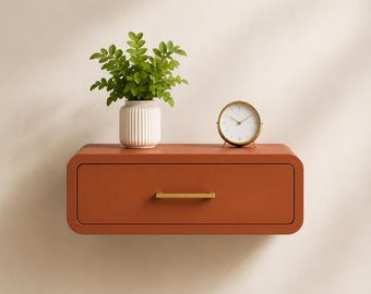 May include: A wall-mounted floating drawer in a matte terracotta finish with a horizontal gold handle. On top sits a white ribbed ceramic vase containing a green leafy plant and a round gold-rimmed analog clock with a white face.