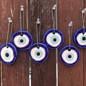 Könnte beinhalten: Sechs blaue und weiße Glücksbringer in Form eines bösen Auges mit silbernen Ketten, die an Haken an einer Holzwand hängen.
