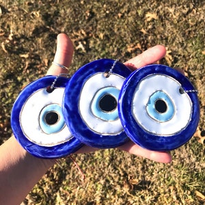 May include: Three blue and white ceramic evil eye charms with gold accents. The charms are round and have a white center with a blue eye in the middle. The charms are hanging from silver chains.