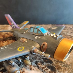 May include: A black and gold model airplane with a blue and white checkered pattern on the side. The plane has a yellow circle with a black outline on the wing and a blue tinted canopy. The plane has a gold tip on the tail and a purple and white stripe on the tail.